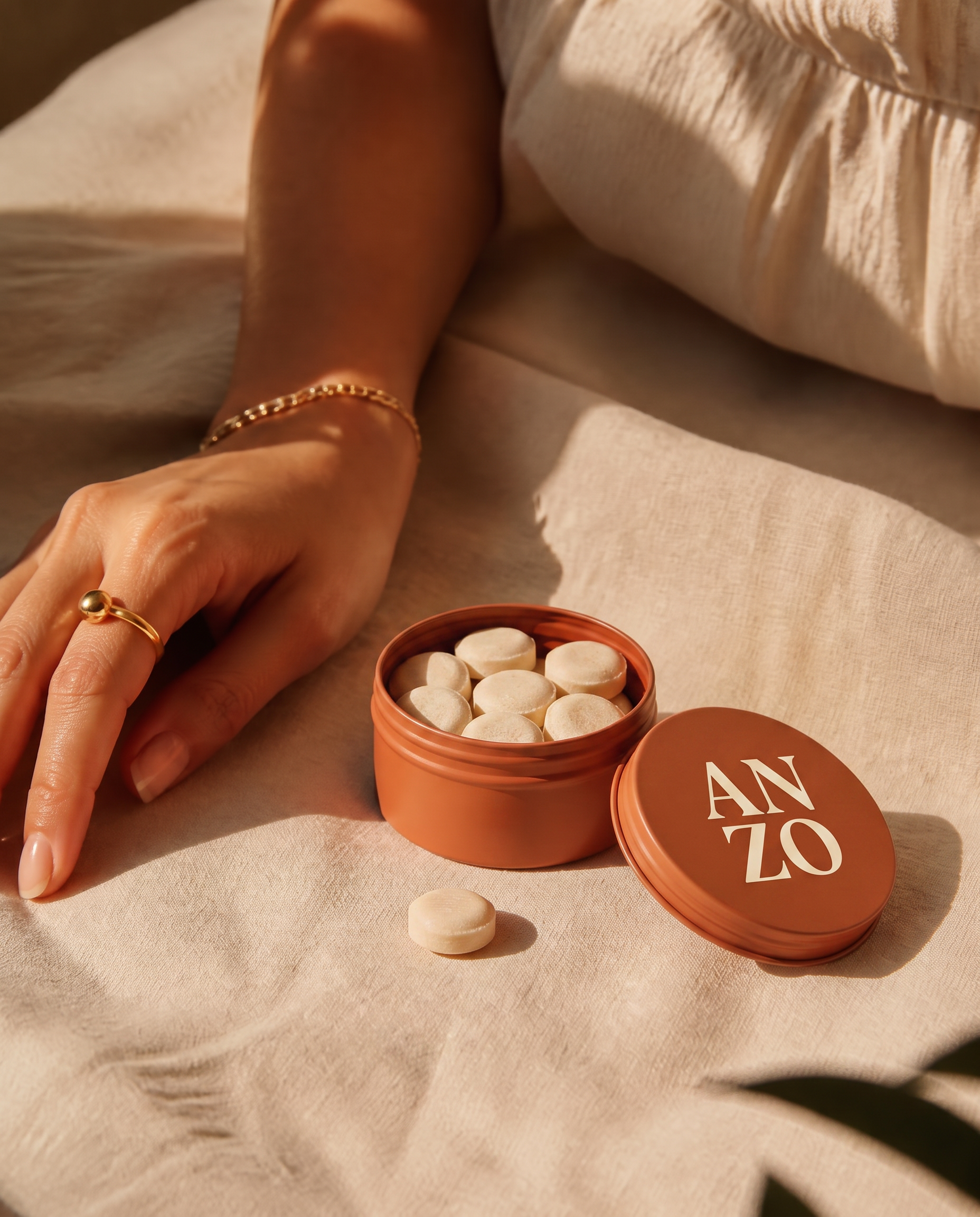 A hand resting beside an open orange ANZO tin and a single pastille on warm cream linen
