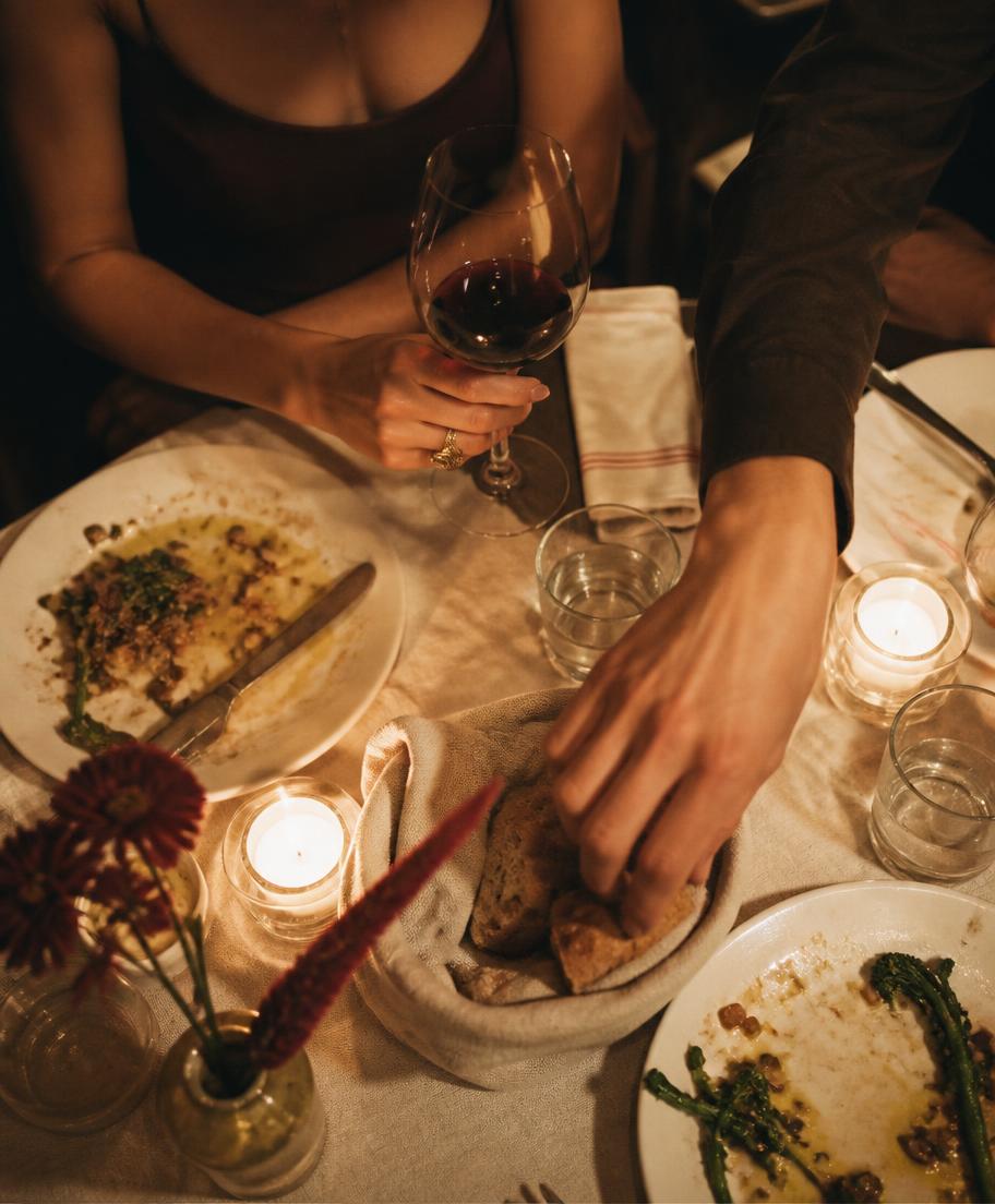 Candlelit dinner table with a hand loosely holding a wine glass and another hand reaching for bread among half-finished plates